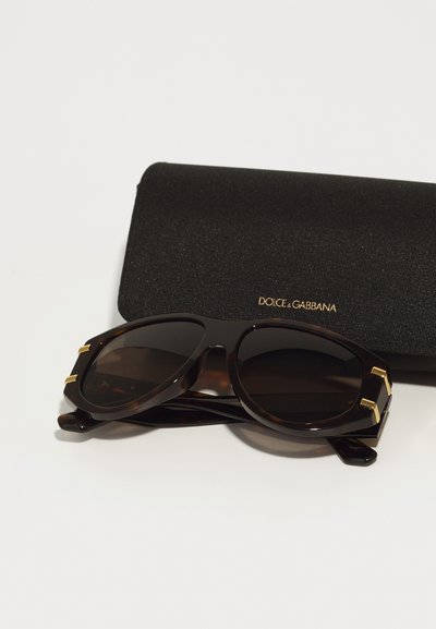 Kilpkonnakuju prillid tumedate läätsedega, kuldsed détailid templitel, asetatud tekstuurse musta karbi kõrvale, millel on "DOLCE & GABBANA" logo.