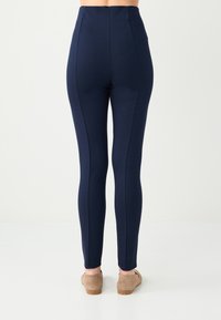 Leggings bleu marine taille haute en tissu extensible, avec détails sur les coutures latérales et silhouette ajustée. Des chaussures en daim sont portées.