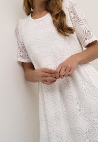 Valkoisista pitsimekko, jossa on kukkakuvio, lyhyet hihat ja pyöreä kaula-auko. Pehmeä tekstuuri ja löysä malli, jossa on kerätty vyötärö.