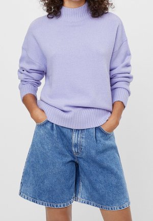 Person trägt einen hellvioletten, gerippten Pullover mit hochgekrempelten Ärmeln und lockere blaue Jeans-Shorts, die die Hände in den Taschen hat.