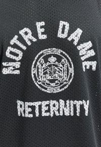 Tissu en mesh noir avec texte blanc usé « NOTRE DAME » courbé au-dessus d'un emblème circulaire et « RETERNITY » en dessous.