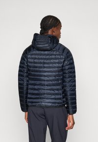 Donkerblauwe pufferjack met capuchon, een gewatteerd design met horizontale strepen en een gladde textuur. Gecombineerd met donkere broek.
