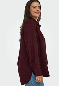 Burgundská košile s dlouhým rukávem, texturovaná, se zapínáním na knoflíky, s bočními rozparky, v kombinaci s světle modrými džíny.