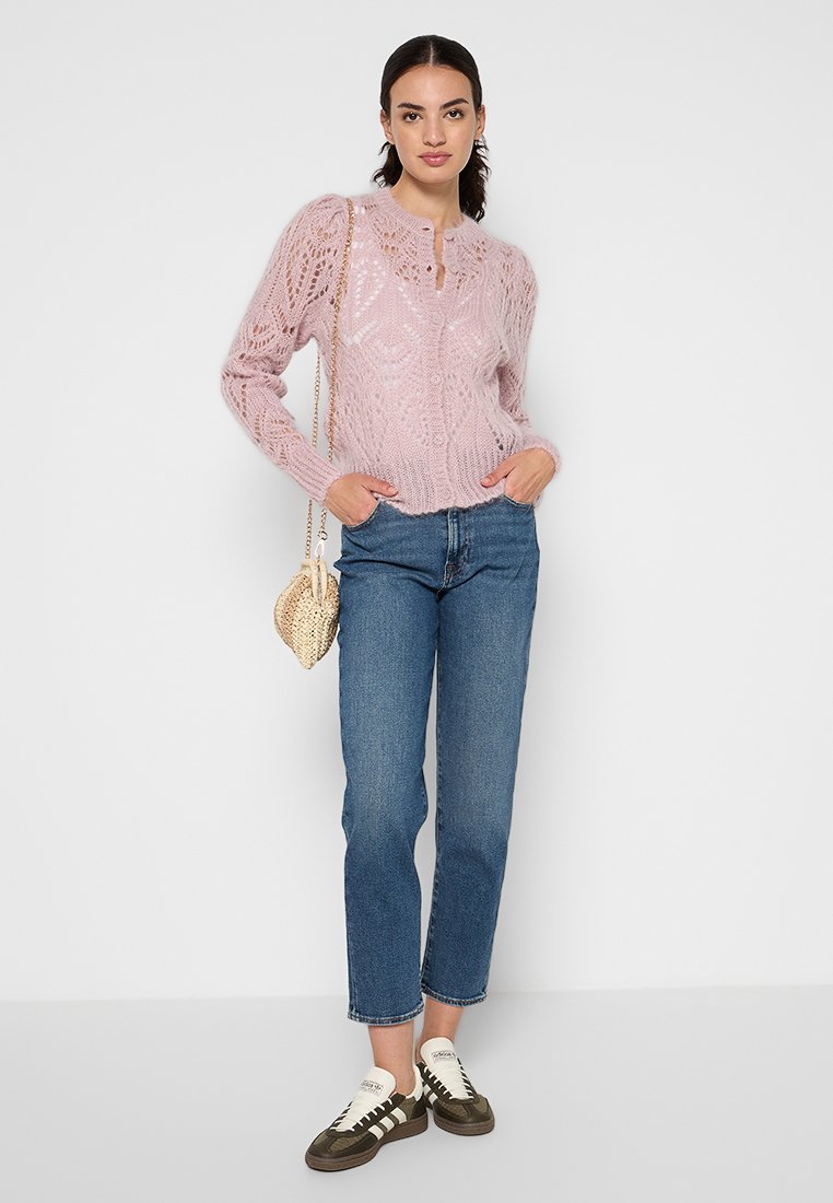 Jasnoróżowy sweter z dzianiny o teksturowanym wzorze, okrągłym dekolcie i zapięciu na guziki, zestawiony z niebieskimi dżinsami i wygodnym obuwiem.