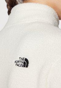 Valge fliisjakke kõrge kraega, millel on must brodeeritud logo The North Face vasakul küljel. Pehme tekstuur ja minimalistlik disain.