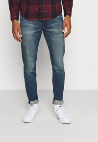 Uomo che indossa jeans blu arrotolati, sneaker bianche e una camicia a quadri rossa e nera, in piedi contro uno sfondo semplice.