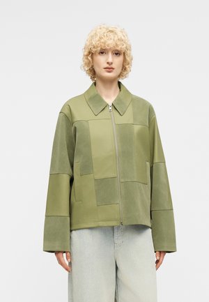 Άτομο με κοντά σγουρά ξανθά μαλλιά που φοράει ένα μπουφάν ζιπ-απ από μπαλώματα σε ελιά πράσινο χρώμα και τζιν με ανοιχτό πλύσιμο, μπροστά σε απλό φόντο.