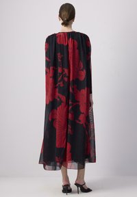 Abito lungo nero fluente con motivo floreale rosso, maniche lunghe, scollatura arricciata e texture trasparente; abbinato a sandali neri con tacco alto.