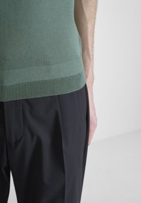 Suéter verde de punto con un dobladillo acanalado, combinado con pantalones negros con pliegues. Enfócate en la textura y el contraste de color entre las prendas.