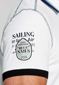 T-shirt bianco in cotone con testo ricamato e un patch con il logo "BLUE SAILS". Orlo a costine neri in contrasto sulle maniche, dettagli in cucitura grigia.