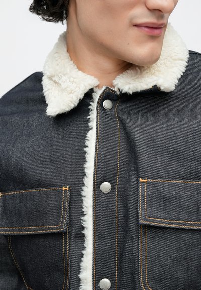 Džínová bunda s krémovým umělým kožíškem kolem límce, stříbrnými knoflíky, oranžovým prošíváním a dvěma předními kapsami na tmavém látkovém pozadí.