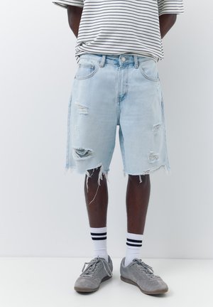 Person trägt hellblaue zerrissene Jeansshorts, weiße gestreifte Socken, graue Sneakers und ein weiß-schwarz gestreiftes Shirt mit den Armen hinter dem Rücken.