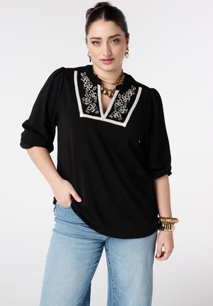 Femme portant un chemisier noir avec une broderie florale blanche sur le col carré et un jean bleu clair, accessoirisée avec des bijoux en or.