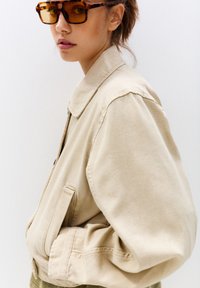 Veste bomber en coton beige avec un col classique, des poches latérales et une coupe décontractée. Complétée par des lunettes de soleil oversize en écaille de tortue.