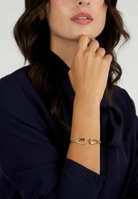 Bracciale in oro con design a goccia, caratterizzato da un accento di cristallo trasparente. Indossato su un polso, abbinato a una blusa blu navy.