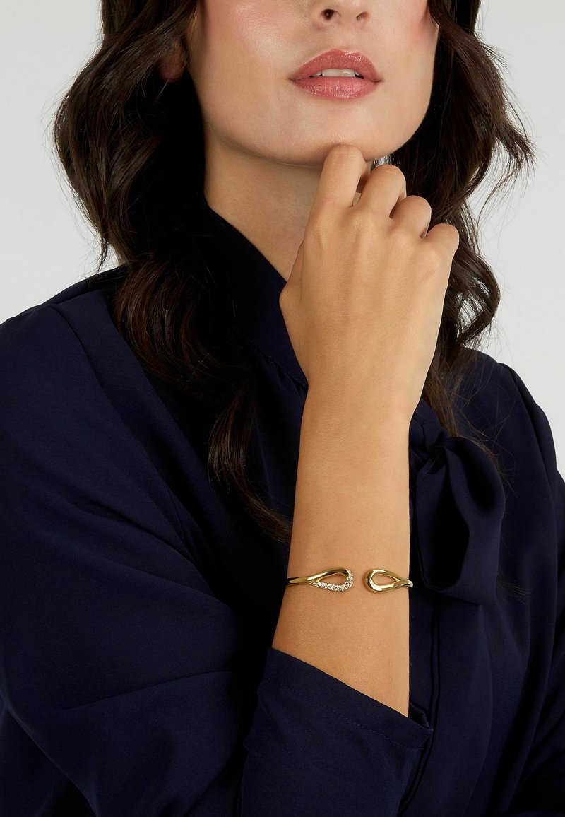 Bracciale in oro con design a goccia, caratterizzato da un accento di cristallo trasparente. Indossato su un polso, abbinato a una blusa blu navy.