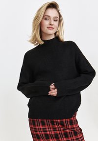 Czarny sweter z dzianiny z wysokim kołnierzem i prążkowanymi mankietami, noszony na czerwonej spódnicy w kratę. Miękka tekstura i luźny krój.