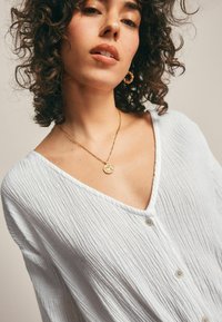 Blouse blanche texturée avec uncol V, dotée d'une fermeture à boutons et de plis subtils. Pendentif de collier en or visible avec un design circulaire.
