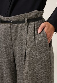 Pantaloni herringbone grigi con vita alta, pieghe e tasche laterali. Abbinati a una cintura nera e una blusa scura. Tessuto testurizzato.