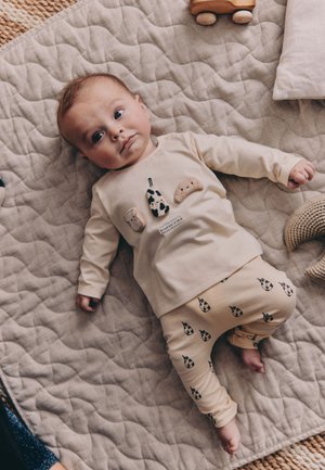 Baba bézs színű, steppelt takarón fekszik, krémszínű ruházatot visel, amelyen ételek témájú minták vannak, körülötte fa játék és puha szőtt tárgyak.