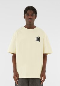 Cream-colored oversized t-shirt with short sleeves, featuring a black graphic on the left chest and a round neckline. Soft cotton material.