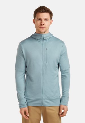 Man draagt een lichtblauwe hoodie met rits en duimgaten en een beige broek, staand tegen een effen witte achtergrond.