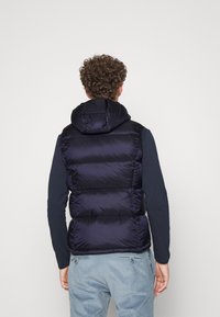 Mann mit lockigem Haar, der eine marineblaue gepolsterte Kapuzenweste über einem Langarmshirt und einer hellblauen Cordhose trägt, und mit dem Rücken zur Kamera steht.