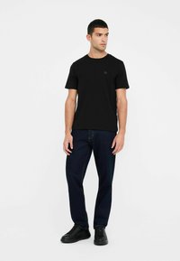 T-shirt nera a maniche corte in una vestibilità rilassata, con una texture liscia e un piccolo logo sul petto, abbinata a jeans blu scuro e scarpe nere.