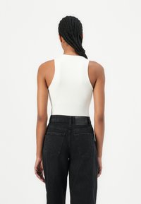 Hvit ermeløs bodysuit kombinert med høyvekkede svarte jeans. Har et rent design, glatt tekstur og subtil merkevare på linningen.