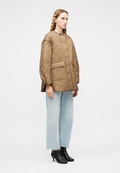 Γυναίκα που φοράει καφέ μπουφάν με επένδυση και μεγάλες τσέπες, ανοιχτό μπλε τζιν με φαρδιά πόδια και μαύρα μποτάκια αστραγάλου, στέκεται μπροστά σε απλό φόντο.