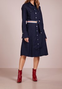 Tummansininen farkkupaita-mekko, jossa on napillinen etuosa, kaksi rintataskua ja monivärinen vyö. Laskostetut hihat lisäävät yksityiskohtia.