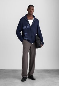 Marinblå stickad zip-up cardigan med grafisk text, matchad med grå byxor och svarta skor. Modellen bär en svart väska.