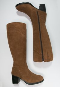 Botas de ante marrón hasta la rodilla con un acabado texturizado, que cuentan con una cremallera lateral y un tacón cuadrado negro para mayor soporte y estabilidad.