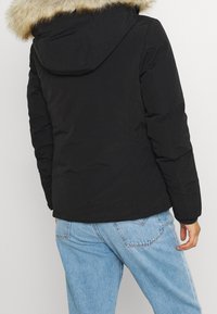 Svart isolerad jacka med avtagbar fuskpälshuva. Designen har en slät yta och åtsittande ärmar, i kombination med ljusblå jeans.