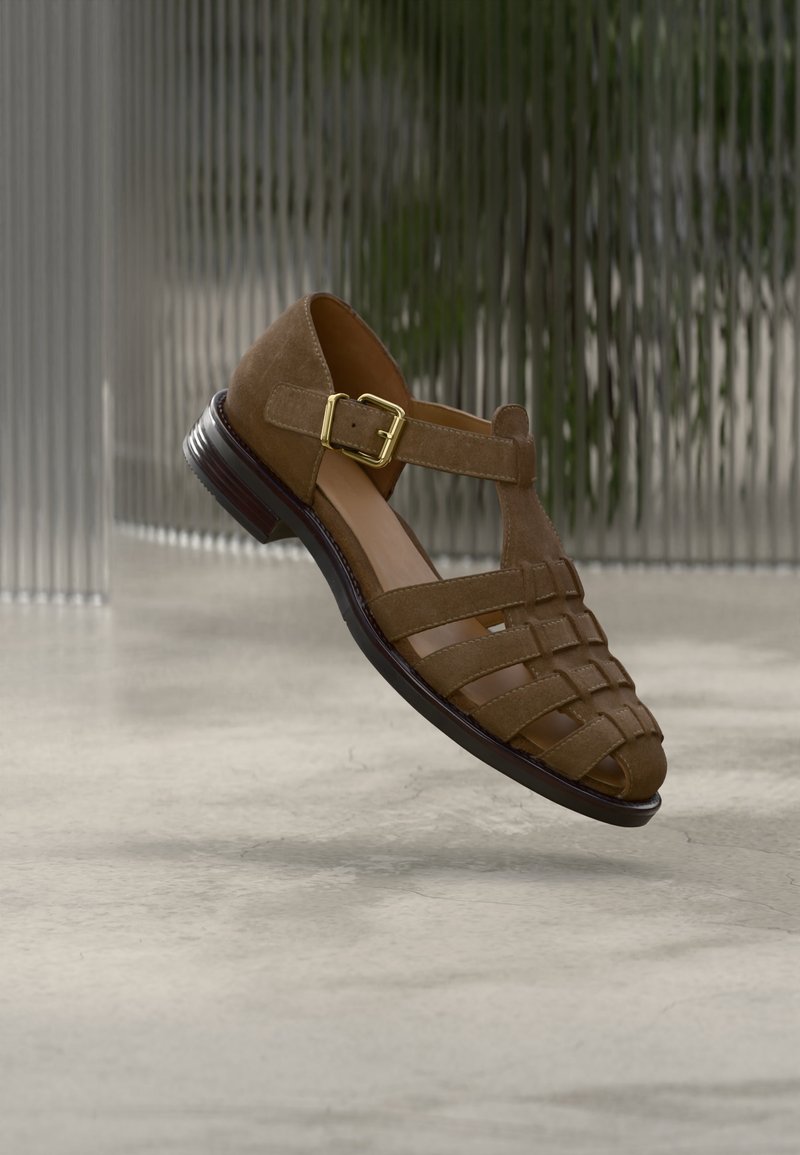 Καφέ δερμάτινο σανδάλι από σουέτ με πλεκτή υφή, χρυσή αγκράφα και χαμηλό τακούνι, σε ουδέτερο φόντο με υφή.