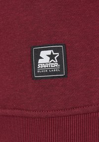 Burgundy színű pulóver anyag bordázott aljjal. Fekete téglalap alakú címke, amelyen fehér "STARTER" felirat és csillaglogó található, kiemelve a "BLACK LABEL" szöveggel.
