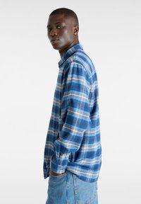 Nuori mies, joka pukeutuu sinivalkoraidalliseen paitaan ja vaaleansinisiin farkkuihin, seisoo sivuttain kädet taskuissa tasaisella taustalla.