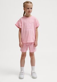 Rosa bomullströja med volanger på ärmarna och en logotyp på framsidan, tillsammans med matchande rosa shorts och vita sneakers.