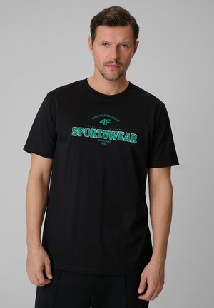 Mężczyzna z krótkimi włosami i brodą, ubrany w czarną koszulkę z zielonym napisem "SPORTSWEAR" na szarym tle.