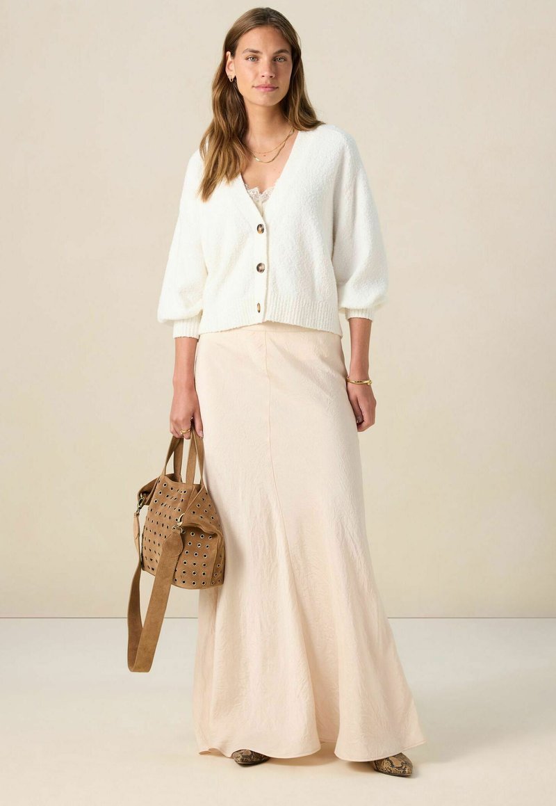 Vrouw in een witte geknoopte cardigan, een beige satijnen maxirok, slangenhuid schoenen en een tan suède handtas met metalen oogjes.