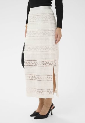 Moteris dėvi baltą nėriniais puoštą midi sijoną su šonine skeltuke, juodą ilgomis rankovėmis palaidinę, juodus slingback stiletus ir laiko juodą rankinę.