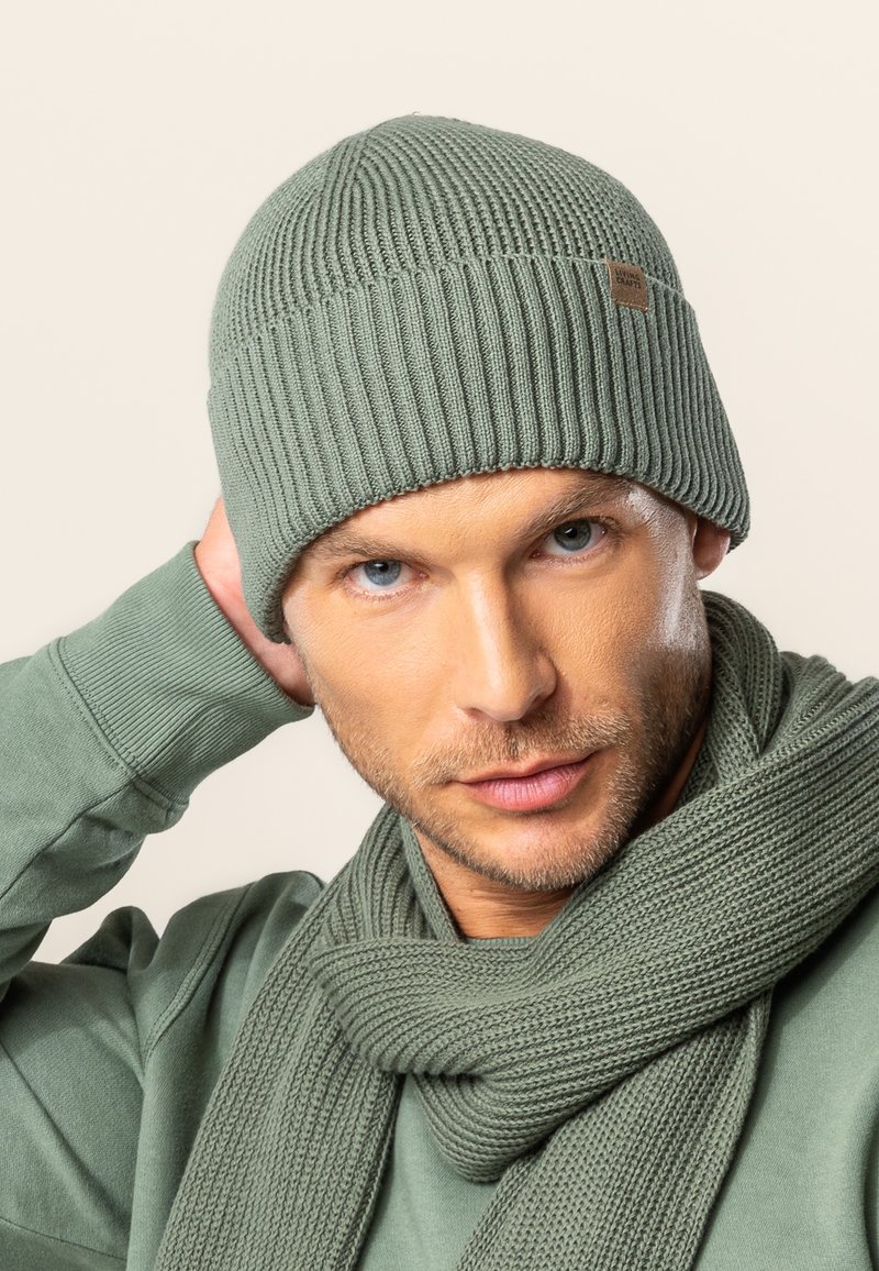 Knitted green beanie with a ribbed texture and a small label, paired with a matching scarf, displayed against a neutral background.