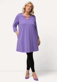 Abito tunica viola con maniche a tre quarti, caratterizzato da un collo a V e dettagli incrociati; abbinato a leggings neri e tacchi viola.