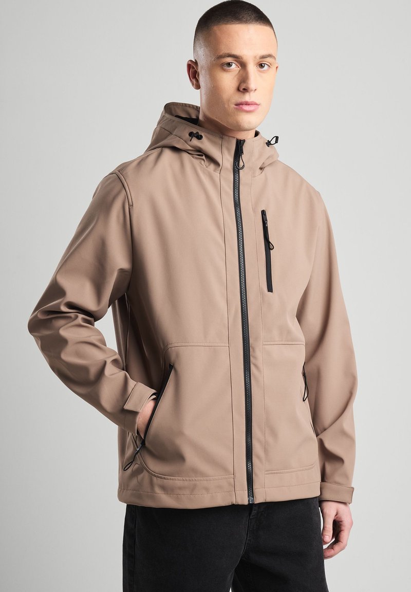 Beige softshell jacket with a hood, black zipper, and side pockets. Features a smooth texture and adjustable Drawstrings at the hood.