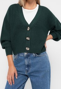 Cardigan verde a maglia con scollo a V, dotato di tre grandi bottoni chiari e maniche a tre quarti. Abbinato a jeans in denim blu.