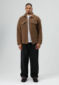 Brown fuzzy jacket with a collar and two chest pockets, worn over a white t-shirt, paired with loose black jeans and black shoes.