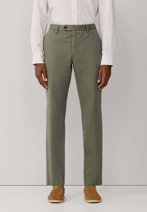 Hombre con pantalones rectos color verde oliva, camisa beige a rayas con botones metida por dentro y zapatos de ante marrón claro sin cordones.