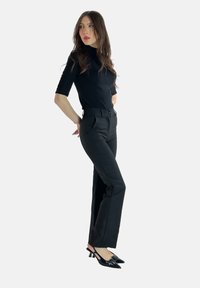 Top nero aderente con maniche corte abbinato a pantaloni neri a vita alta con pieghe frontali. Modella che indossa tacchi a punta neri.