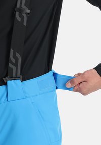 Blaue wasserdichte Hose verfügt über verstellbare Träger und einen Klettverschlussbund. Der Stoff sieht glatt aus und hat eine lockere Passform mit практичischem Design.