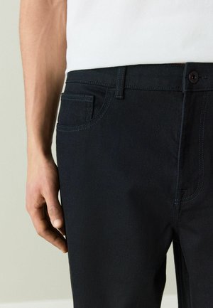 Mann trägt schwarze Denim-Jeans und ein weißes Hemd, die Hand entspannt an der Seite, zeigt Details der Vordertasche und des Knopfverschlusses.